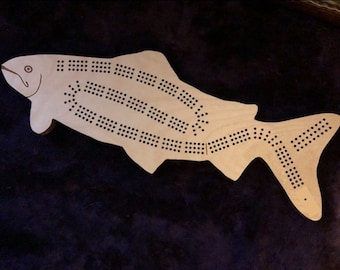 May include: A wooden fish-shaped cribbage board with a natural wood finish. The board has a series of holes for pegs and a scoring track.