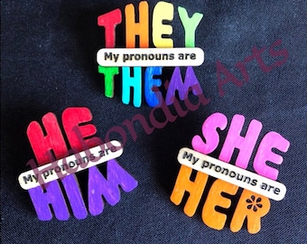 May include: Four wooden pronoun pins with rainbow colors. The pins read "They/Them", "He/Him", "She/Her", and "My pronouns are".