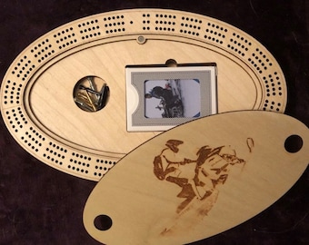 May include: A wooden cribbage board with a hinged lid. The lid features a laser-engraved image of a person riding a snowmobile. The board has a compartment for storing pegs and a card holder.