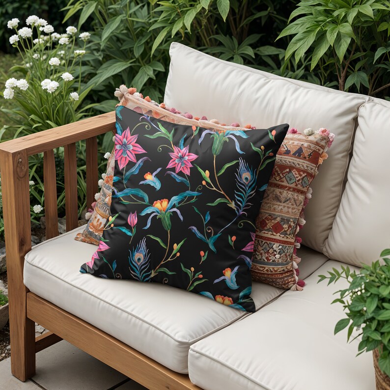 May include: A black and white cushion with a floral pattern featuring pink, blue, and yellow flowers and peacock feathers. The cushion is on a white sofa with a wooden frame.