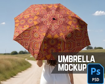 Könnte beinhalten: Ein offener Regenschirm mit einem detaillierten, symmetrischen Muster in Rot-, Orange- und Gelbtönen. Der Regenschirm wird von einer Person gehalten, die auf einem Feldweg geht. Der Text "UMBRELLA MOCKUP" ist sichtbar.