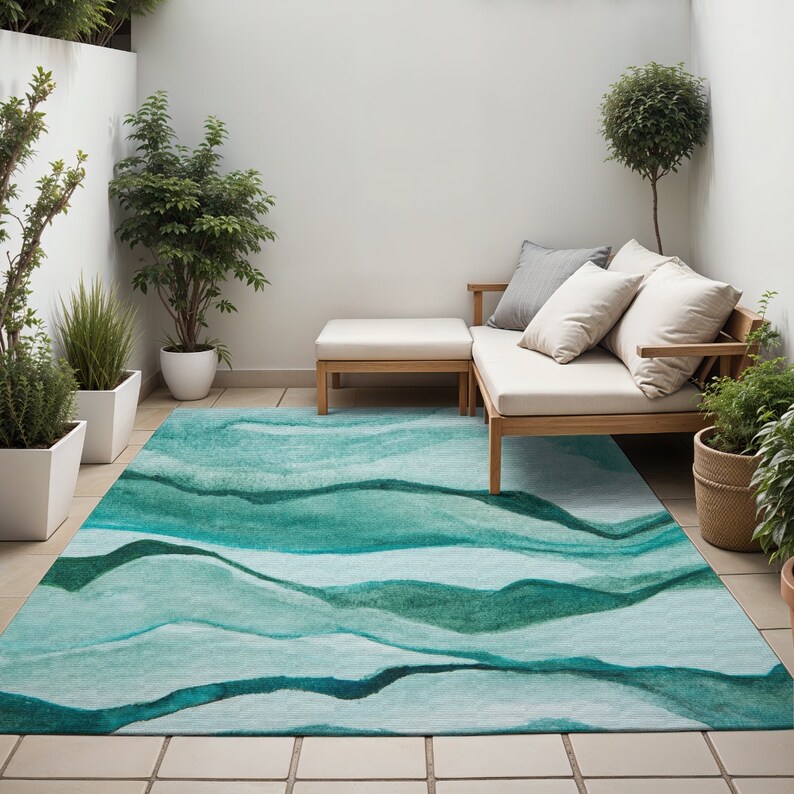 May include: A teal and green abstract patterned outdoor rug with a light blue background. The rug is placed in a patio setting with a white couch and a wooden coffee table.