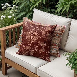 May include: Two throw pillows on a light brown wooden outdoor sofa. The front pillow is a deep red with a paisley pattern in a lighter shade of red. The back pillow is a multi-coloured patterned fabric with tassels.