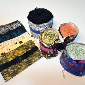 Könnte beinhalten: Sortiment an bunten Garnen in verschiedenen Farben und Mustern, aufbewahrt in Garnbehältern aus Stoff mit Riemen. Die Garnbehälter sind aus verschiedenen Stoffen mit floralen, geometrischen und abstrakten Mustern.