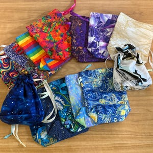 May include: A collection of fabric drawstring bags in various sizes and patterns. Designs include rainbow stripes, swirling patterns, a starry night sky, and floral prints. Some bags have ribbon or cord closures.