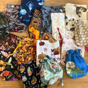 Peut inclure: Un assortiment de pochettes à cordon avec divers motifs, notamment des ratons laveurs, des chats, des colibris et des citrouilles. Les pochettes sont en tissu et ont des cordons de couleurs différentes.