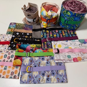 May include: A collection of colorful fabric yarn holders with various patterns and designs. Each holder has a strap with a buckle to secure the yarn. The holders are made of different fabrics, including floral, geometric, and abstract patterns.