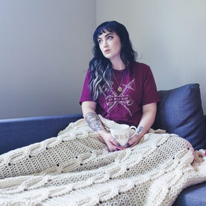 May include: A woman sits on a couch with a white crocheted blanket. She is wearing a maroon t-shirt with the text "We Will Rise" and a gold necklace. She is holding a white mug with the text "Mrs." in gold lettering.