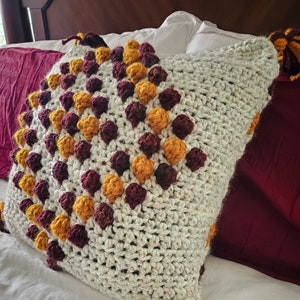 May include: A white crocheted pillow with a textured surface and a geometric pattern of burgundy and mustard yellow pom-poms. The pillow has long burgundy and mustard yellow tassels.