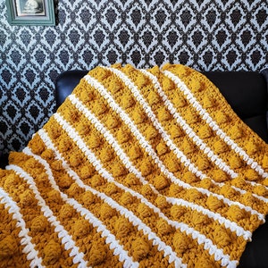 May include: A yellow and white striped crocheted throw blanket with a textured, puffy design. The blanket is folded and draped over a black couch.