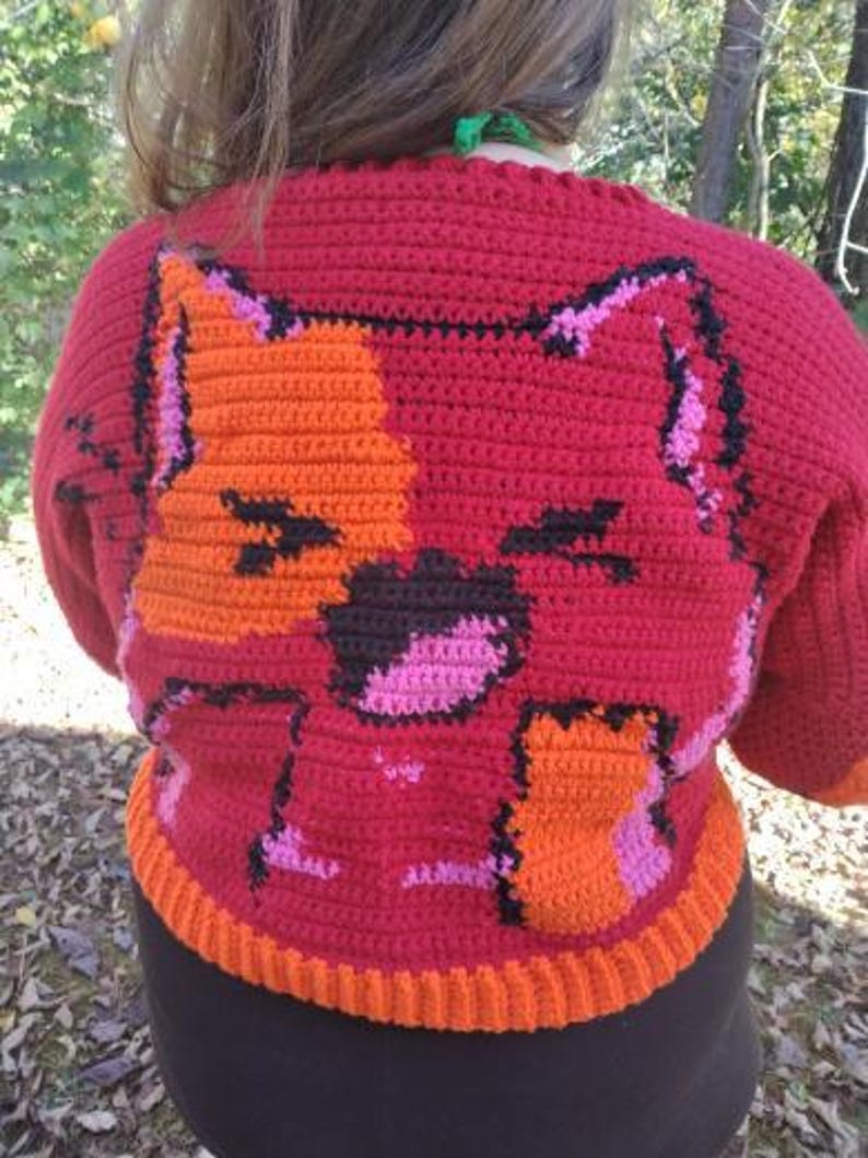 May include: A red crocheted jumper with an orange and pink cat design. The jumper has a ribbed hem and cuffs.