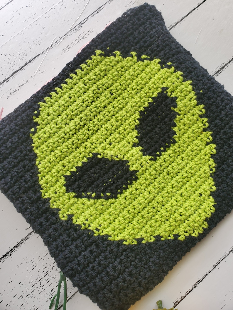 May include: A black and green crocheted pouch with an alien face design. The pouch is made with a textured stitch and has a pink zipper.