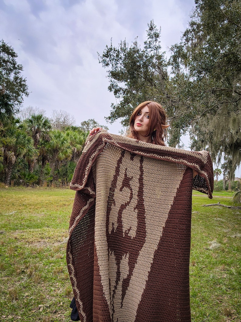 May include: A woman wearing a brown and beige crocheted blanket with a geometric pattern. The blanket is draped over her shoulders and she is holding it up with her hands. She is standing in a grassy field with trees in the background.