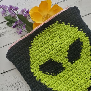 May include: A black crocheted zippered pouch with a green alien face design. The pouch is lying on a white wooden surface with a yellow flower and purple flowers in the background.