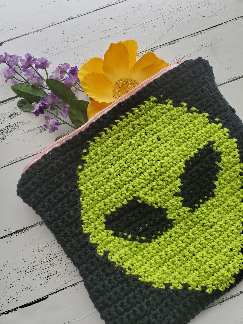 May include: A black crocheted zippered pouch with a green alien face design. The pouch is lying on a white wooden surface with a yellow flower and purple flowers in the background.