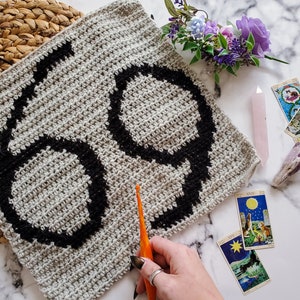 May include: A crocheted square with the number "69" in black yarn on a gray background. The square is being worked on with an orange crochet hook. There are tarot cards and crystals on a white marble surface.