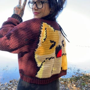 May include: A maroon crocheted cardigan with a cartoon cat design on the back. The cat is yellow, brown, and white with a red mouth.