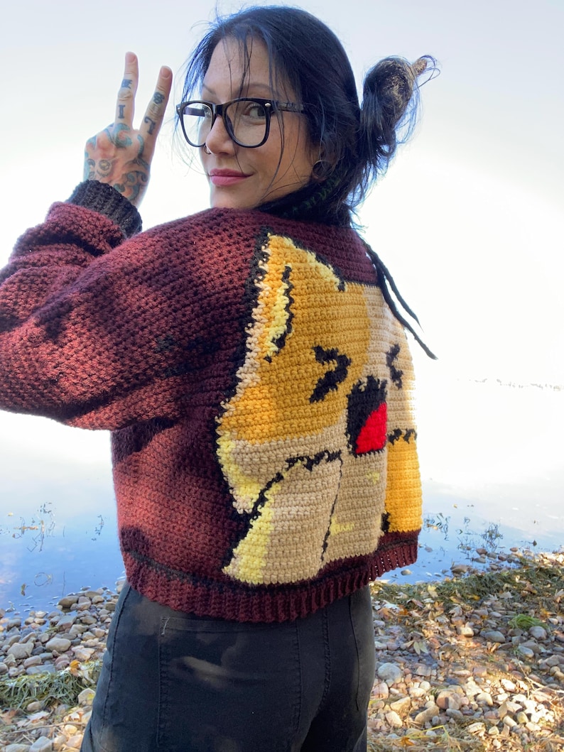 May include: A maroon crocheted cardigan with a cartoon cat design on the back. The cat is yellow, brown, and white with a red mouth.
