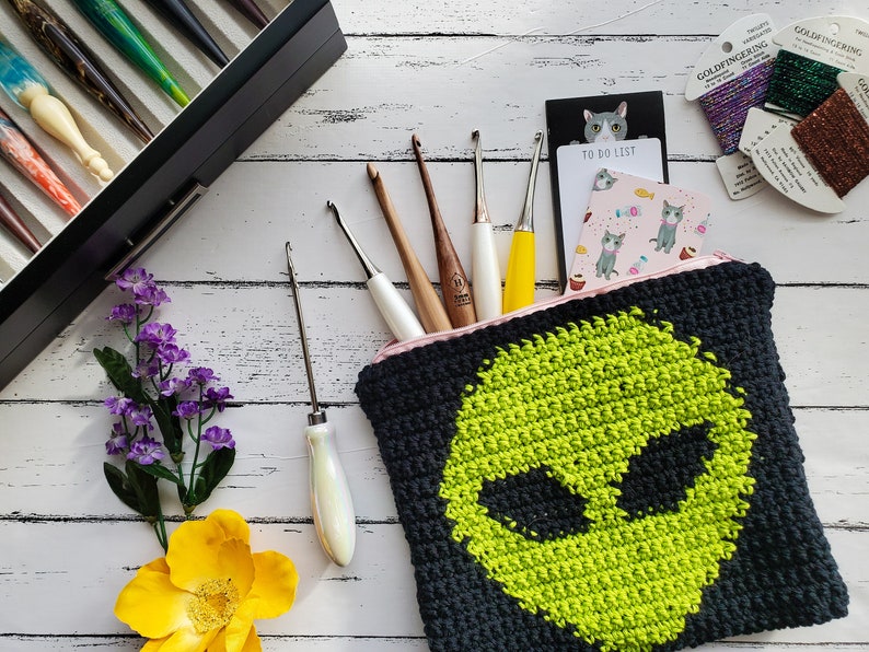 May include: A black and green crocheted zippered pouch with an alien face design. The pouch is filled with crochet hooks and yarn.