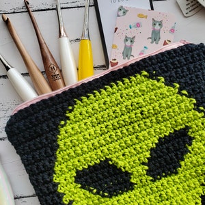 May include: A black and green crocheted zippered pouch with a graphic of an alien face. The pouch is lying on a white wooden surface with crochet hooks and other craft supplies.