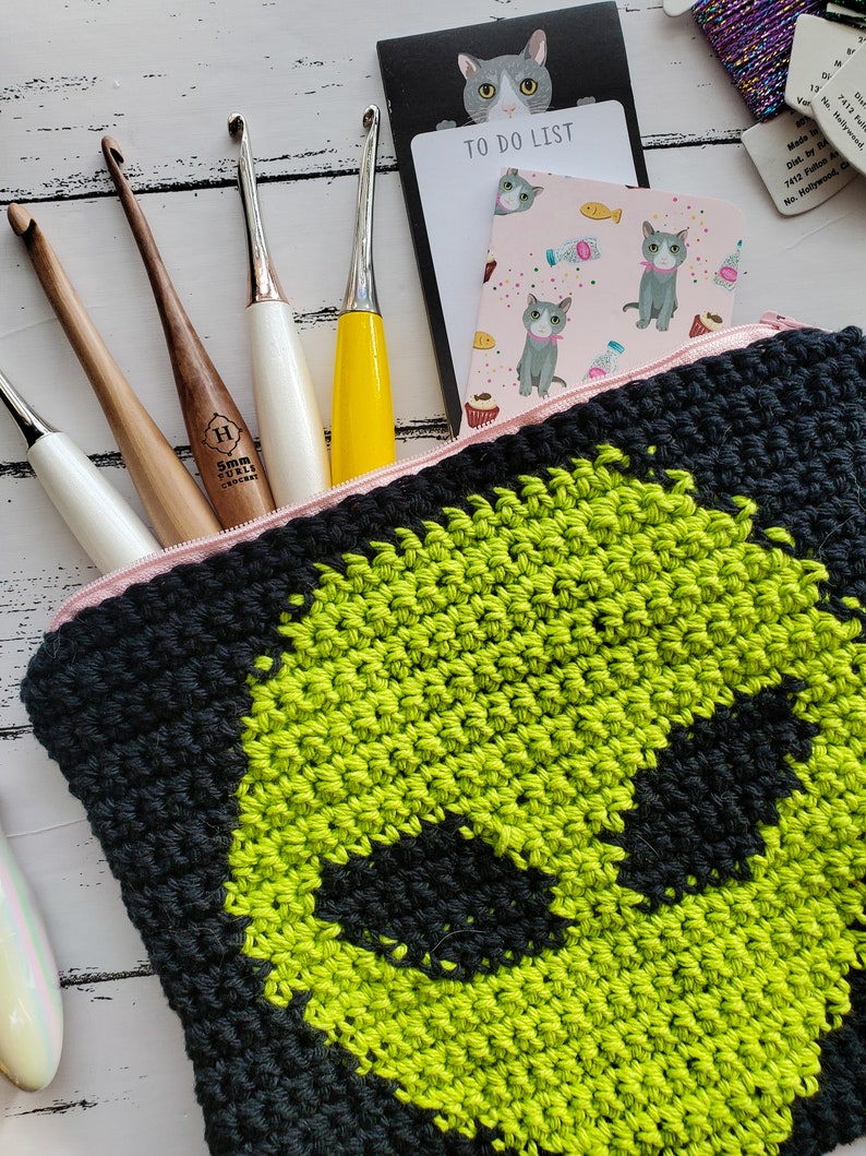 May include: A black and green crocheted zippered pouch with a graphic of an alien face. The pouch is lying on a white wooden surface with crochet hooks and other craft supplies.