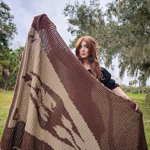 May include: A brown and beige knitted blanket with a geometric pattern. The blanket is held up by a person in a field.