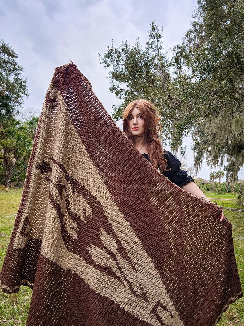 May include: A brown and beige knitted blanket with a geometric pattern. The blanket is held up by a person in a field.