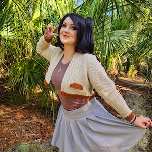 May include: A woman wearing a beige crocheted cropped cardigan with brown accents, a grey skirt, and black tights. She is standing in a wooded area with green plants.