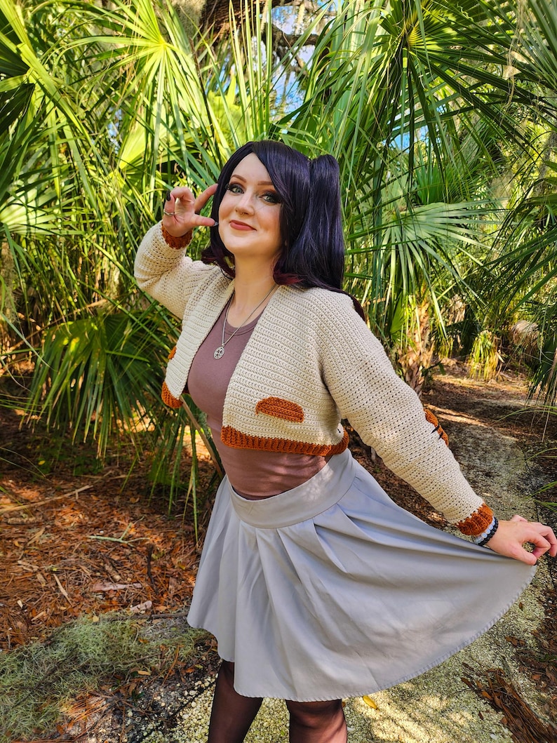 May include: A woman wearing a beige crocheted cropped cardigan with brown accents, a grey skirt, and black tights. She is standing in a wooded area with green plants.