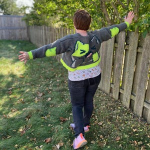 May include: A person wearing a grey and neon green crocheted cardigan with a cat design on the back. The cardigan is cropped and has long sleeves. The person is standing in a grassy area with a wooden fence in the background.