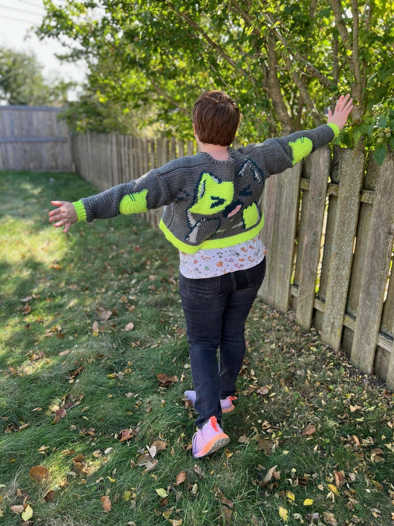 May include: A person wearing a grey and neon green crocheted cardigan with a cat design on the back. The cardigan is cropped and has long sleeves. The person is standing in a grassy area with a wooden fence in the background.