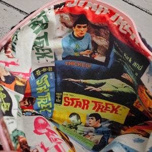 May include: A pink zippered pouch with a white fabric lining featuring a collage of Star Trek comic book covers. The covers feature images of Captain Kirk and the text "Star Trek" and "Tricked!"