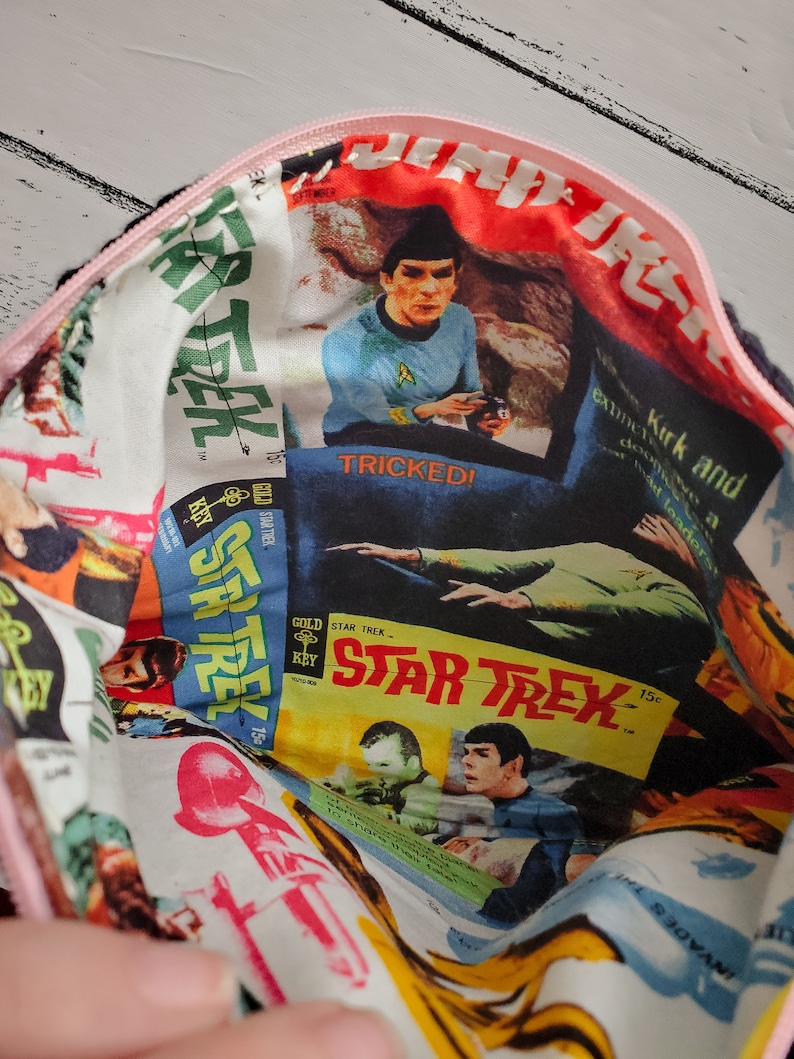 May include: A pink zippered pouch with a white fabric lining featuring a collage of Star Trek comic book covers. The covers feature images of Captain Kirk and the text "Star Trek" and "Tricked!"