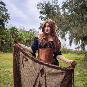 May include: A woman with long brown hair is wearing a brown corset and a black top. She is holding a brown and beige crocheted blanket with a geometric pattern.