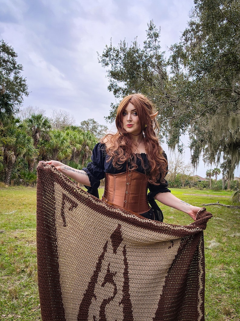 May include: A woman with long brown hair is wearing a brown corset and a black top. She is holding a brown and beige crocheted blanket with a geometric pattern.