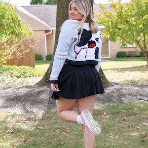 May include: A white cropped cardigan with a black and white cartoon image on the back. The cardigan is worn with a black skirt and pink trainers.