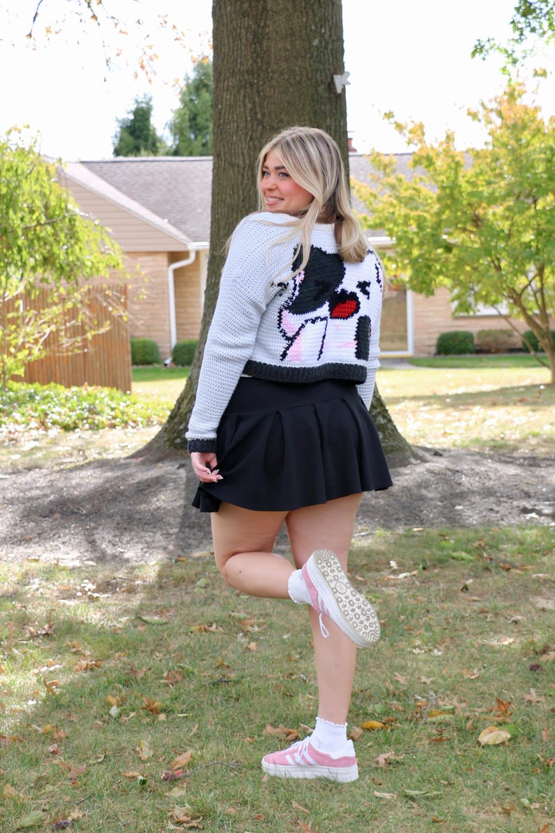May include: A white cropped cardigan with a black and white cartoon image on the back. The cardigan is worn with a black skirt and pink trainers.