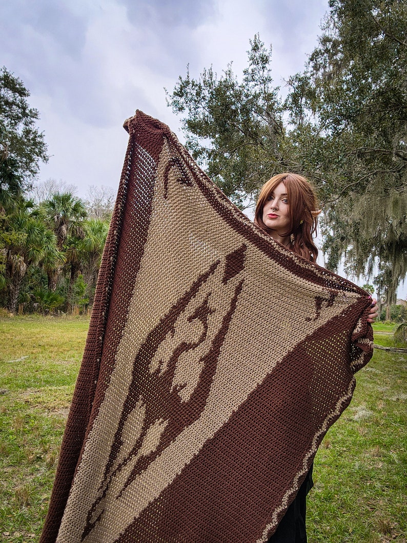 May include: A woman holds a brown and beige knitted blanket with a large, stylized design. The blanket is held up in front of her, and she is looking at the camera.