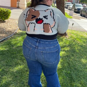 May include: A cropped cardigan with a light blue background and a brown and white cat design on the back. The cardigan has black trim around the edges. The person wearing the cardigan is also wearing blue jeans.
