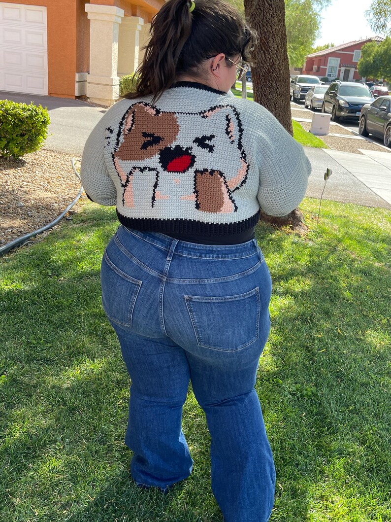 May include: A cropped cardigan with a light blue background and a brown and white cat design on the back. The cardigan has black trim around the edges. The person wearing the cardigan is also wearing blue jeans.