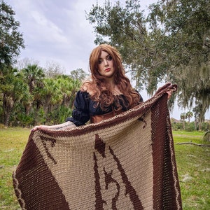 May include: A woman wearing a black off-the-shoulder top and a brown leather corset holds a brown and beige crocheted blanket with a large brown design.