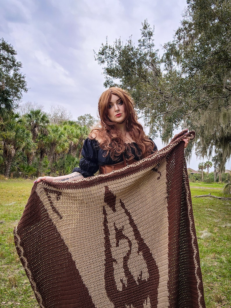May include: A woman wearing a black off-the-shoulder top and a brown leather corset holds a brown and beige crocheted blanket with a large brown design.