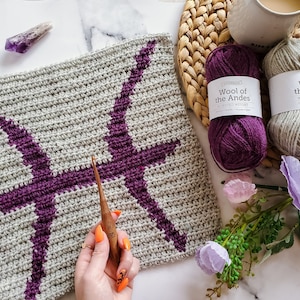 Puede incluir: Un cuadrado tejido a crochet con un signo del zodíaco de Piscis morado en el centro. El cuadrado se está trabajando con un gancho de crochet de madera. Dos ovillos de hilo, uno morado y uno gris, están en el fondo.