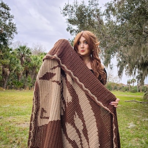 May include: A person wearing a brown and beige crocheted blanket with a leaf pattern. The blanket is held up to the person's face, and they are looking at the camera.