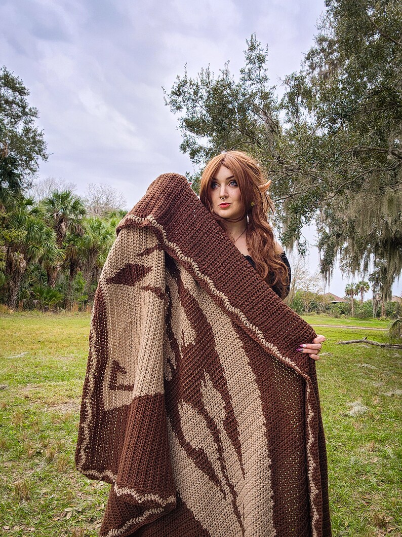 May include: A person wearing a brown and beige crocheted blanket with a leaf pattern. The blanket is held up to the person's face, and they are looking at the camera.