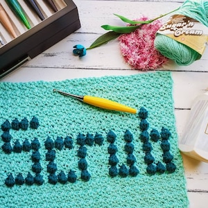 May include: A teal blue crochet dishcloth with the word "Scrub" spelled out in dark blue yarn. The dishcloth is made with a textured stitch and has a raised, puffy look.