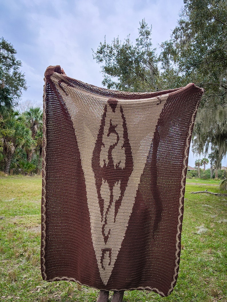 May include: A brown and beige crocheted blanket with a geometric pattern. The blanket is held up by a person's hands, and the background is a grassy field with trees.