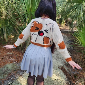 May include: A white crocheted cropped sweater with a brown cat design on the back. The cat has a surprised expression with its mouth open and tongue sticking out. The sweater is worn over a grey pleated skirt.