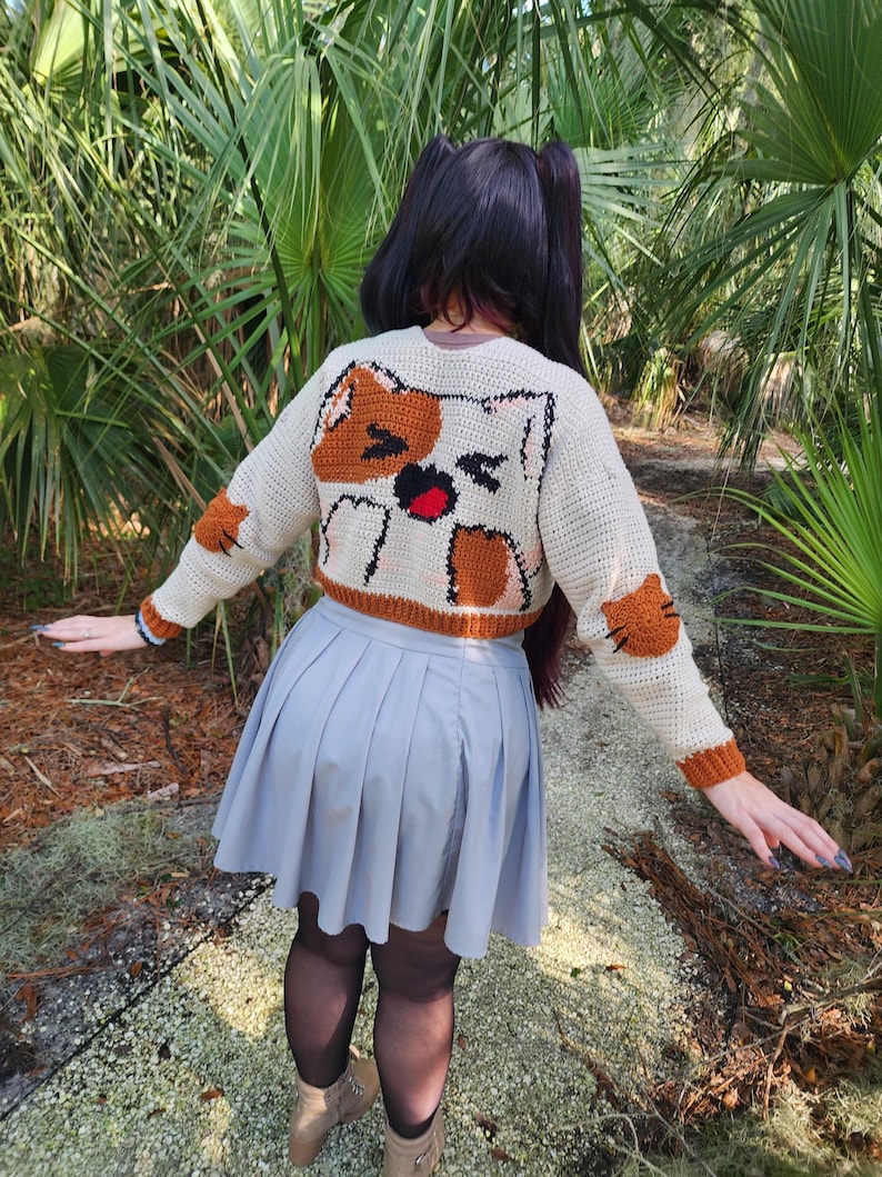 May include: A white crocheted cropped sweater with a brown cat design on the back. The cat has a surprised expression with its mouth open and tongue sticking out. The sweater is worn over a grey pleated skirt.