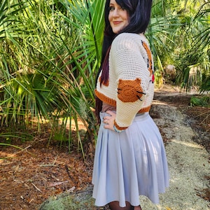 May include: A woman wearing a white and brown crocheted cropped sweater with a cat design. The sweater has a brown cat paw on the sleeve. She is wearing a light blue skirt and black tights.